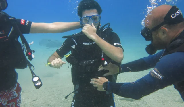 Fethiye Ölüdeniz’de 5 metre tüplü dalış deneyimi sırasında burun kapatarak basınç eşitleyen dalgıç; yanlarda eğitmenler, kumluk zeminde sığ derinlik.
